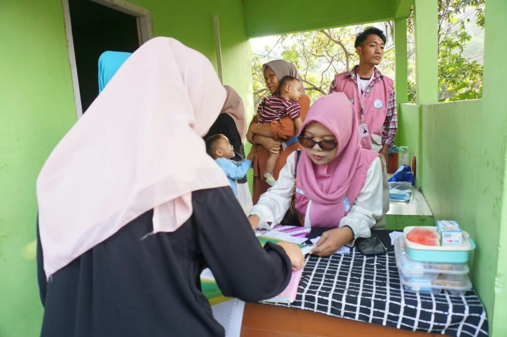 Mahasiswa KKN Universitas Ibnu Chaldun mengenakan rompi krem merah muda sedang melayani pendaftaran ibu-ibu dan balita di meja Posyandu.